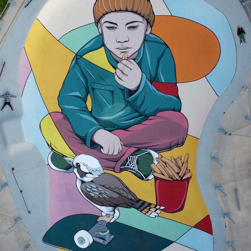 Large-scale mural of a child in a beanie sitting cross-legged with chips, beside a kookaburra perched on a skateboard, painted on a skate park surface.