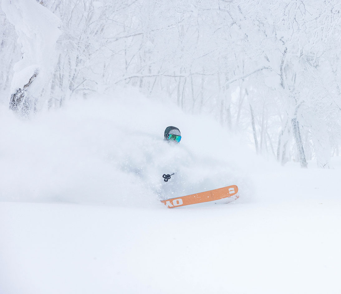 Mike-Shankster-Armada-Kodama-Ski-Artwork-Logo-skier-V7. Skier riding through deep powder with only their goggles and orange ski tips visible in heavy snow.