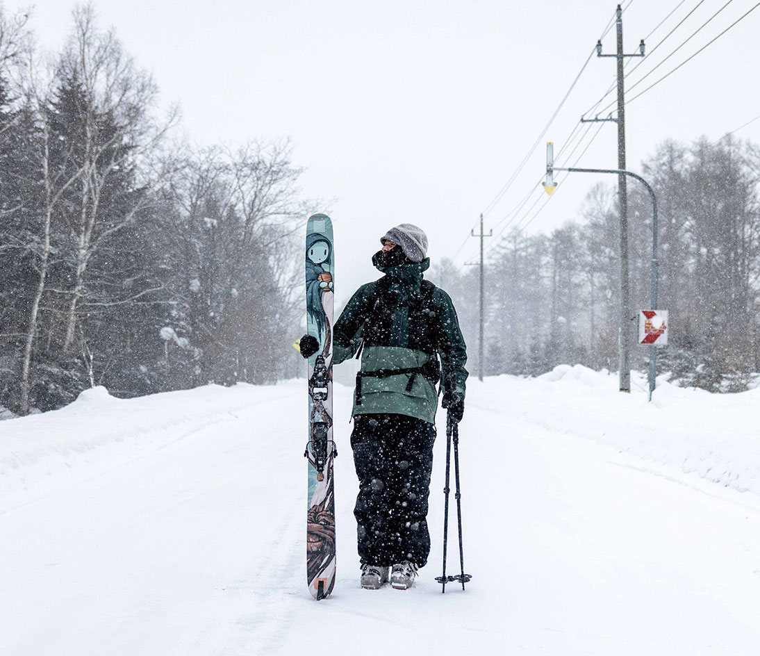 Mike-Shankster-Armada-Kodama-Ski-Artwork-V7 Person standing on a snowy road holding ARV JJ-UL skis featuring Mike Shankster’s Kodama artwork.