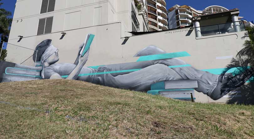 Large-scale Brisbane mural at South Bank depicting a reclining figure reading, painted in a realistic grayscale style with turquoise graphic elements, by Mike Shankster.