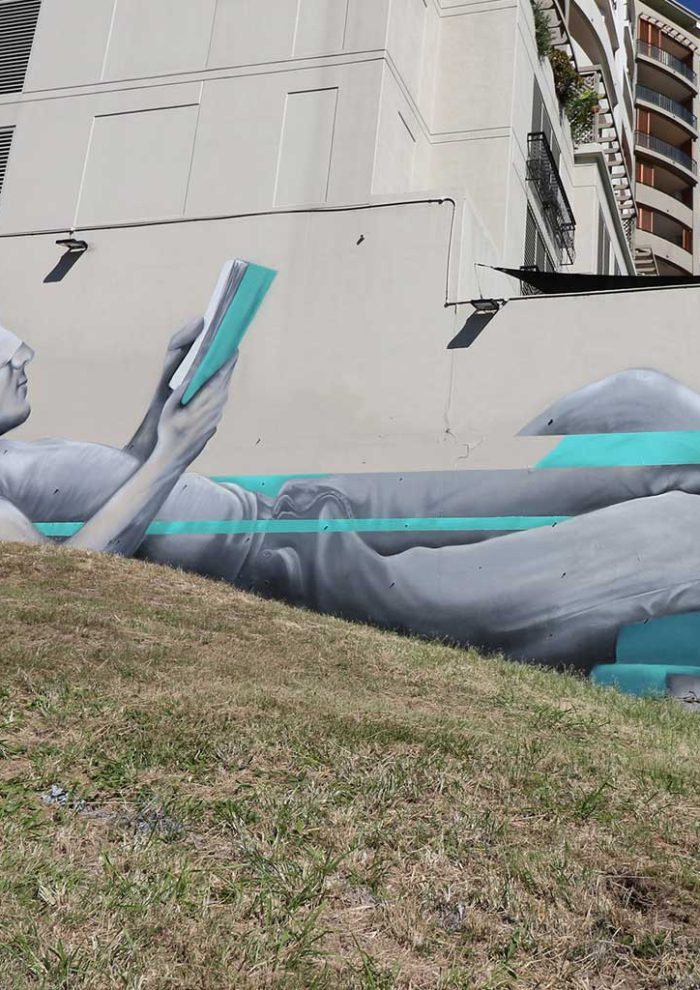 Large-scale Brisbane mural at South Bank depicting a reclining figure reading, painted in a realistic grayscale style with turquoise graphic elements, by Mike Shankster.