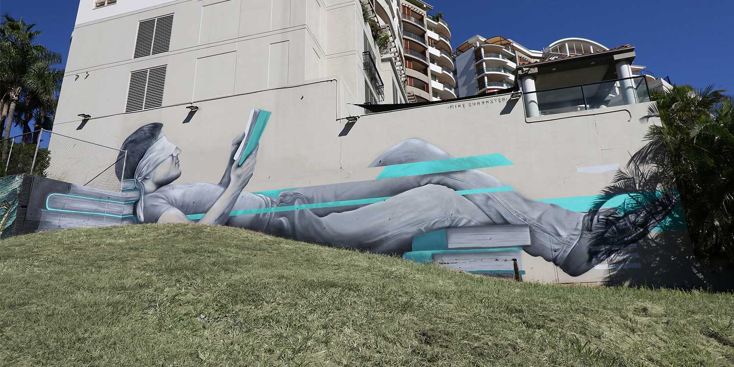Southbank mural by Mike Shankster featuring a large-scale monochrome figure reclining and reading a book, with turquoise graphic accents painted across a retaining wall in Brisbane