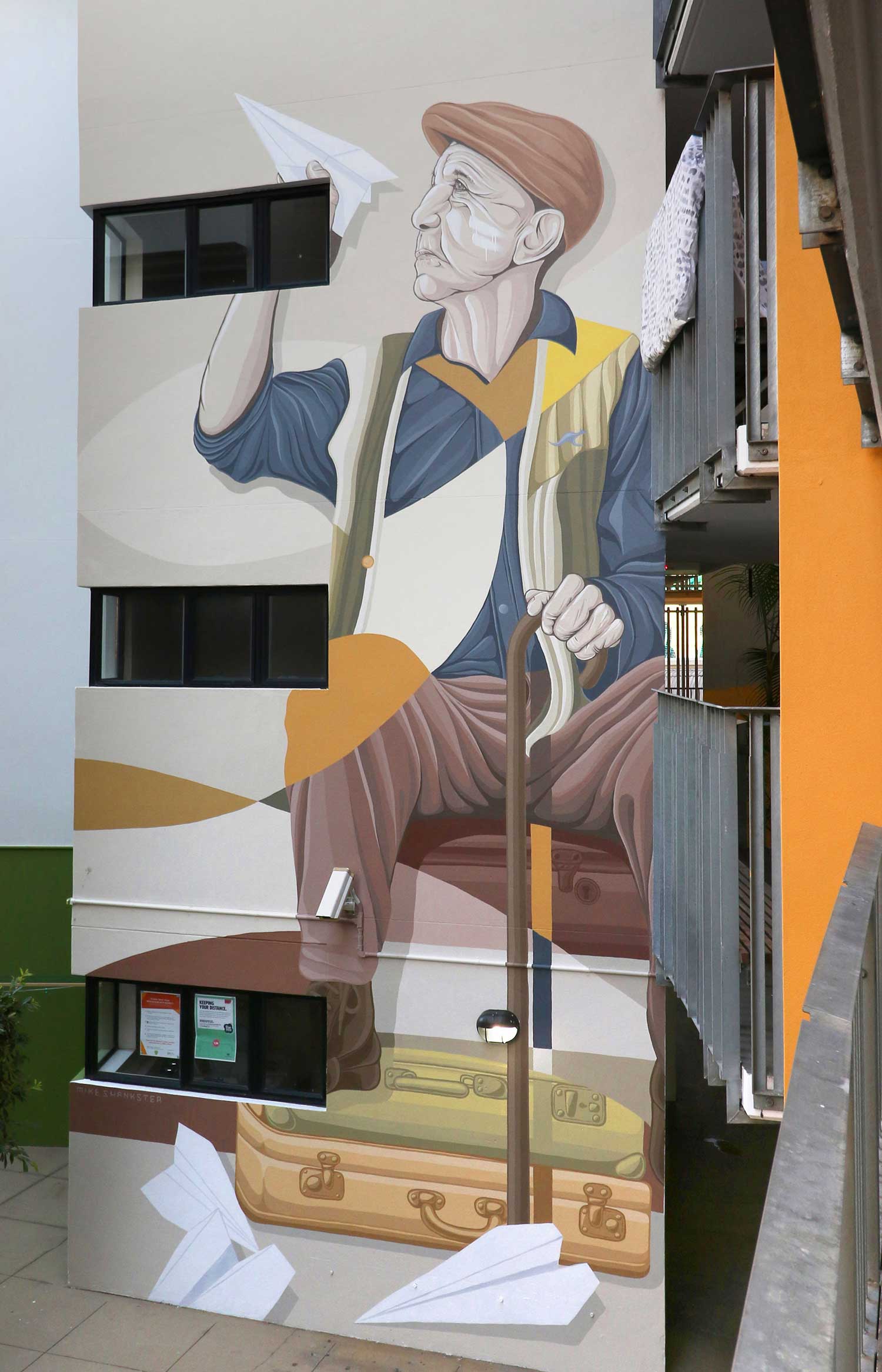 Large-scale mural of an elderly man holding a paper plane, seated on stacked suitcases, Warry Street, Brisbane, by Mike Shankster
