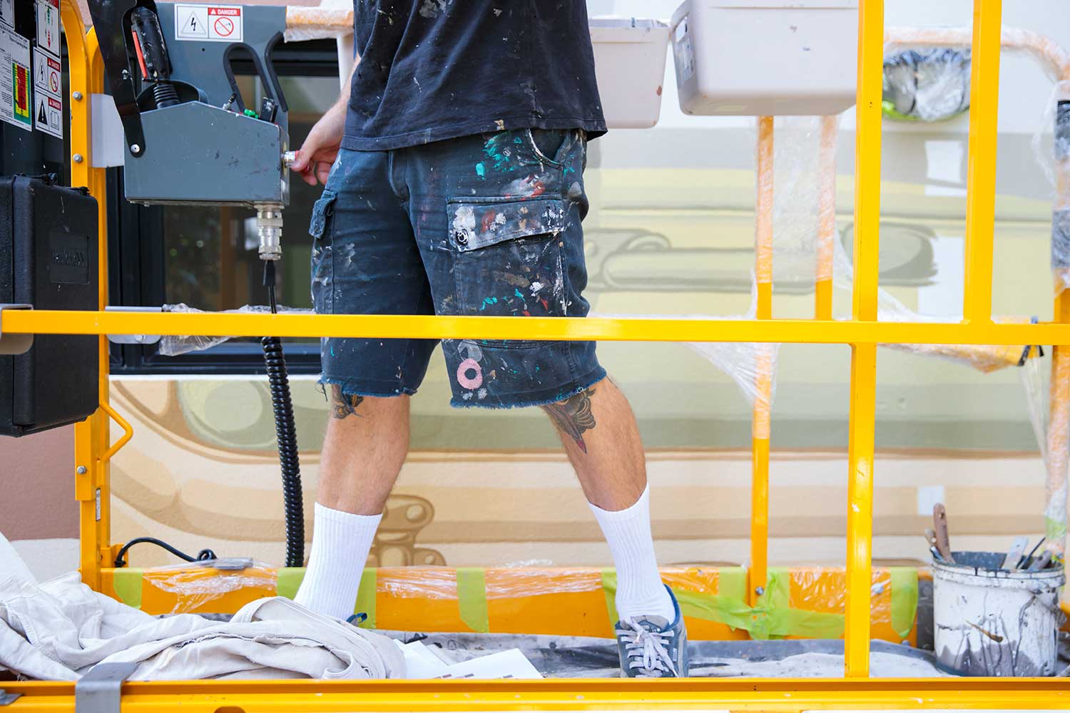 Artist standing on a yellow scissor lift while painting the Warry Street mural in Brisbane Title: Behind the scenes, painting Warry Street mural Brisbane
