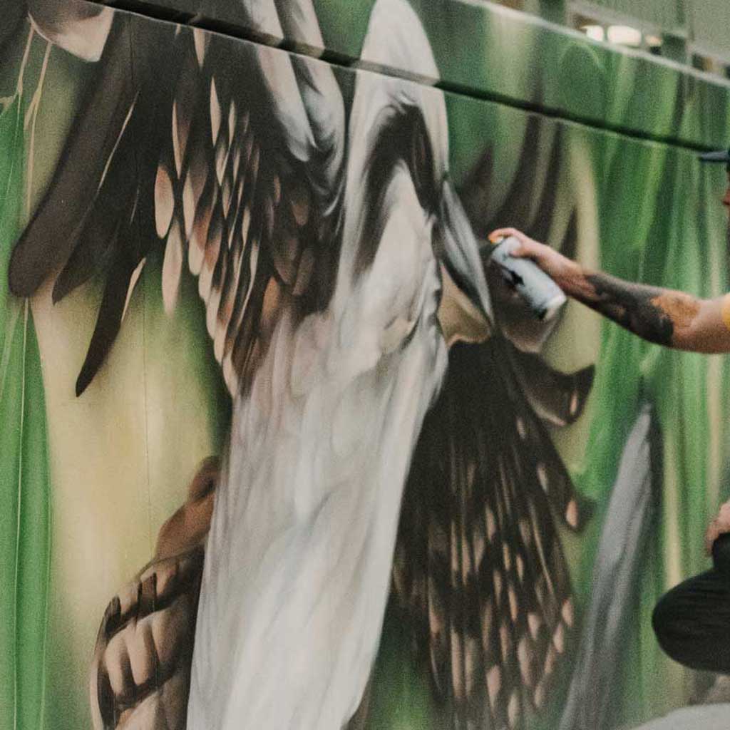Artist painting a large bird mural at Jindabyne Skate Park, with detailed feathers and green tones in the background.