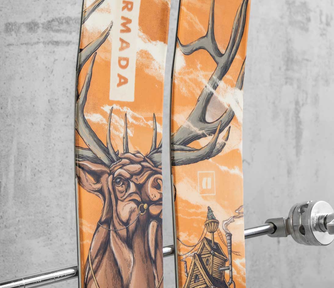 Angled view of Armada skis featuring hand-drawn elk and cabin artwork in warm orange and cream tones.