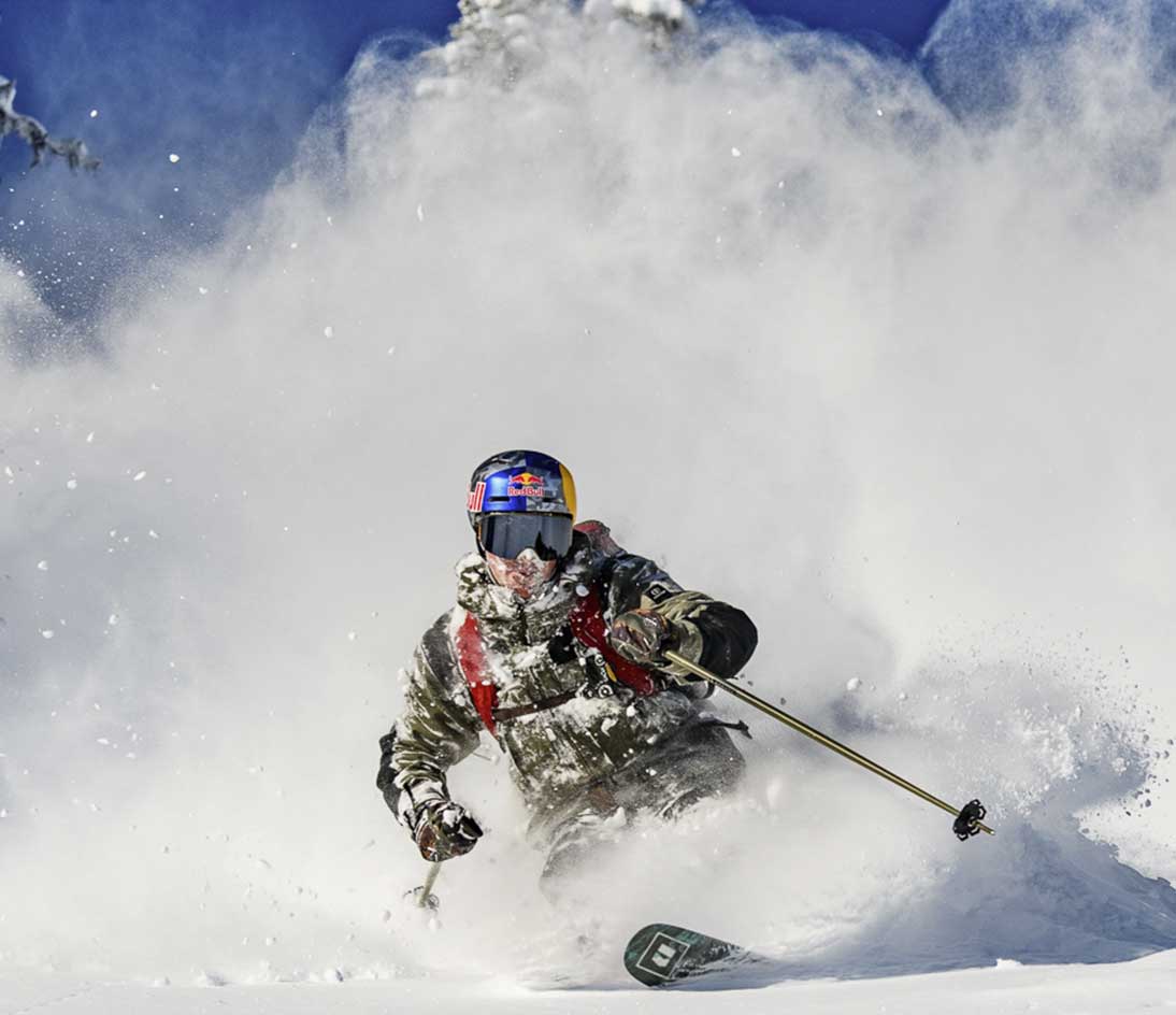Skier riding through deep powder snow on Armada skis in a mountain setting, with snow spray surrounding the rider.