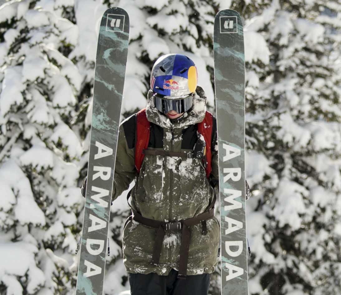 Skier standing in front of snow-covered trees holding a pair of Armada skis upright in the snow.