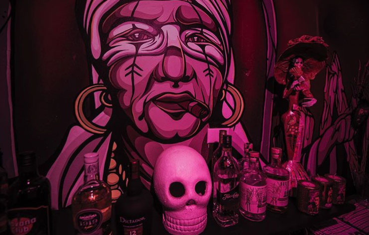 Pink-toned mural portrait of a woman with earrings and a cigar, with bottles and a skull displayed in the foreground.