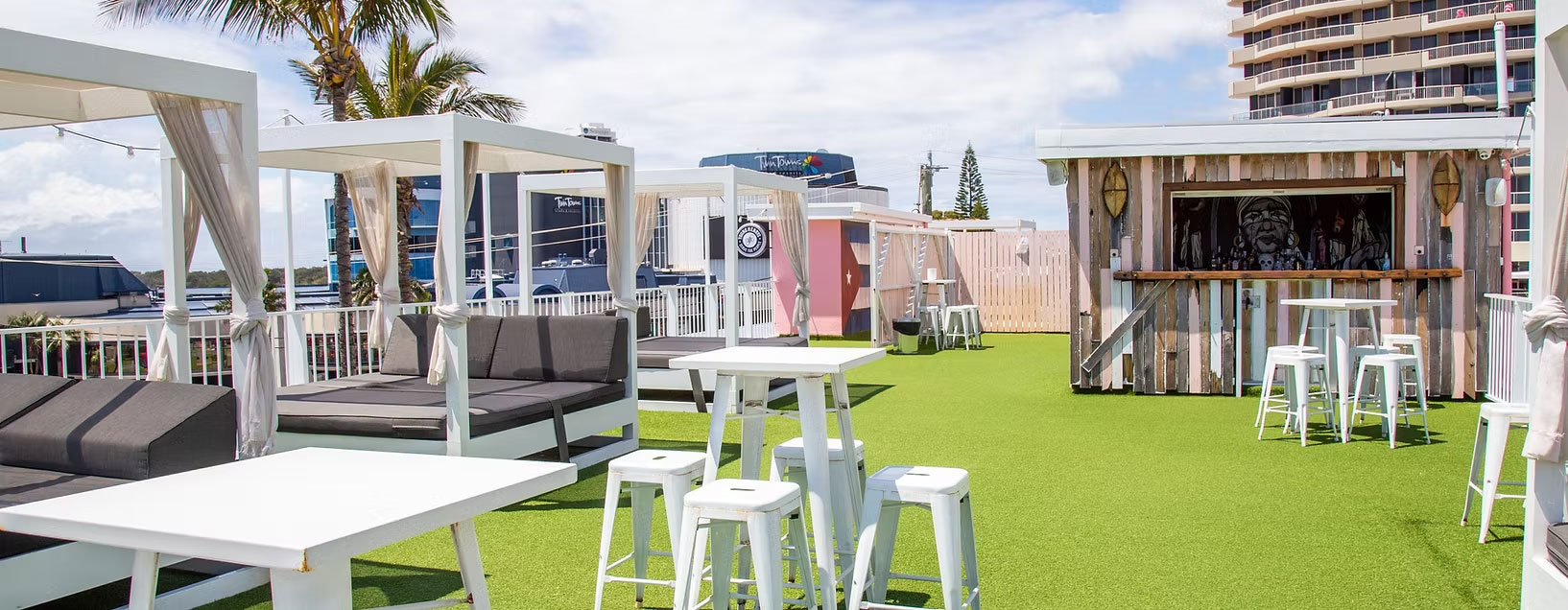 Rooftop bar area at The Pink Hotel in Coolangatta, with white cabanas, tables, stools, and a mural visible behind the bar.