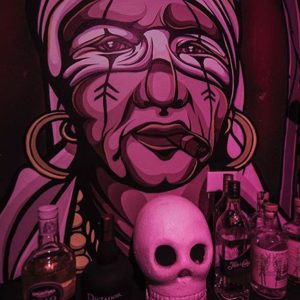 Pink-toned mural portrait of a woman with earrings and a cigar, with bottles and a skull displayed in the foreground.