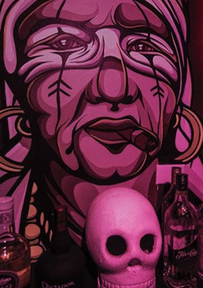Pink-toned mural portrait of a woman with earrings and a cigar, with bottles and a skull displayed in the foreground.