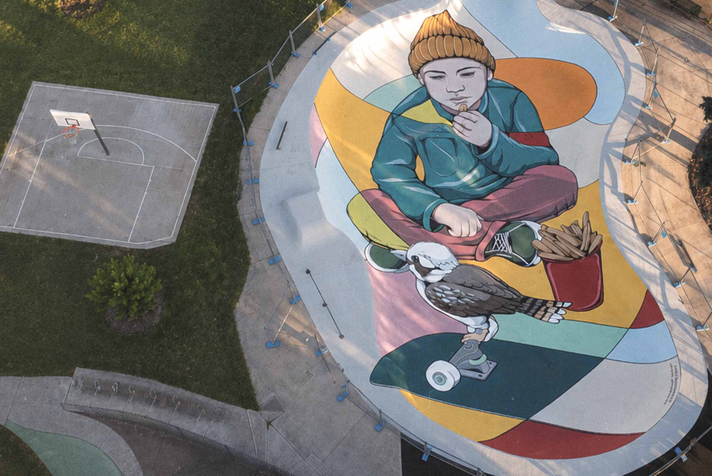 Drone view of a large skate park mural in Queanbeyan featuring a seated child, cockatoo, skateboard and colourful abstract shapes.