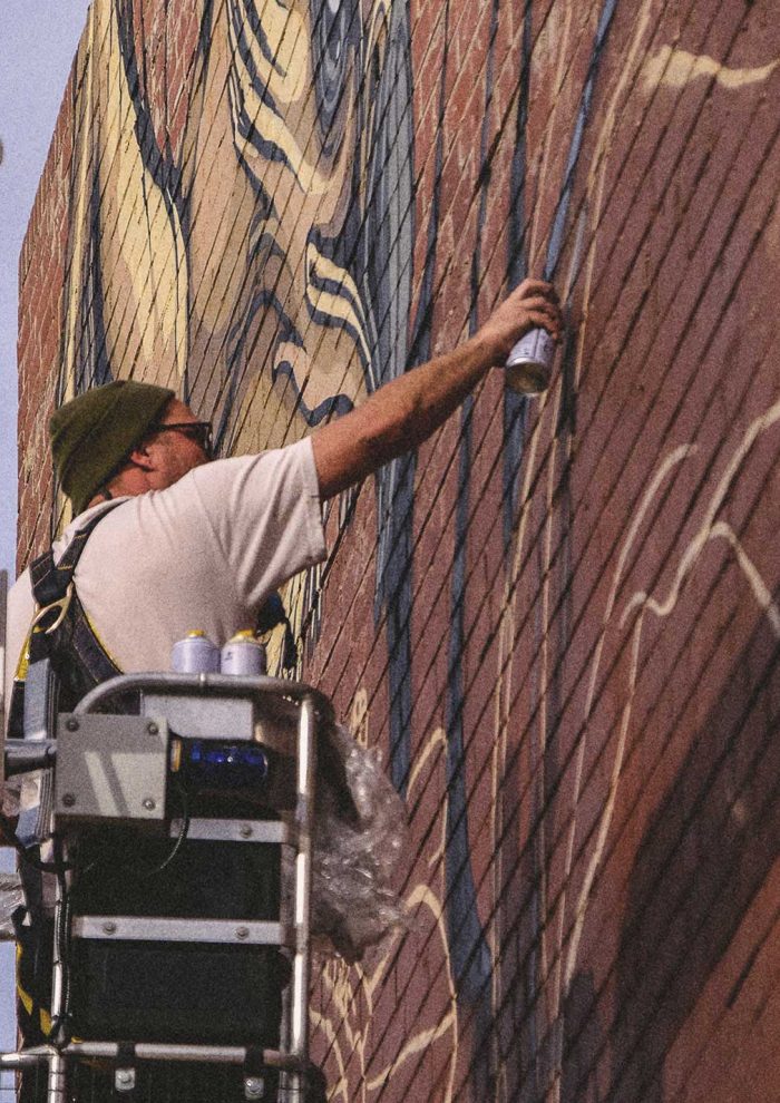 Mike Shankster painting a large-scale brick wall mural from an elevated work platform, using aerosol paint on an outdoor commercial wall.