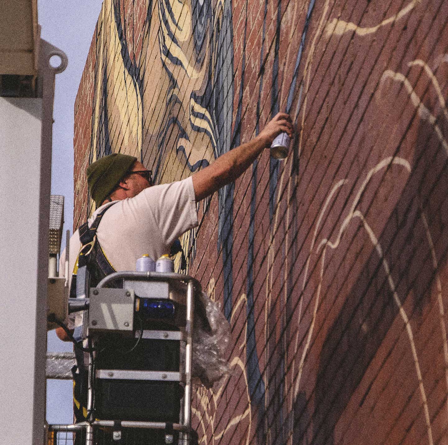 Mike Shankster painting a large-scale brick wall mural from an elevated work platform, using aerosol paint on an outdoor commercial wall.